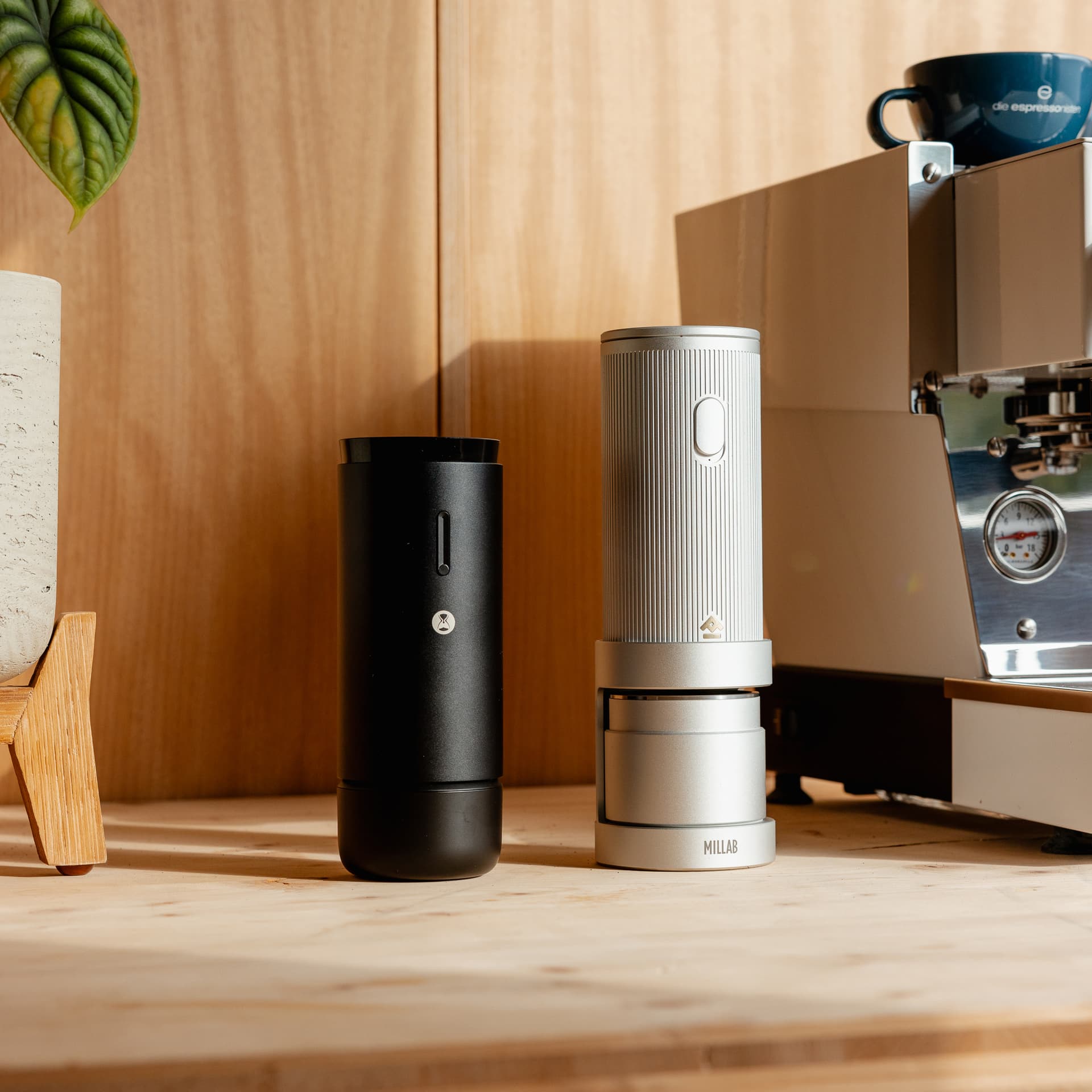 Zwei portable Timemore Handmühlen (schwarz und silber) neben Espressomaschine auf Holzfläche, Millab-Logo sichtbar