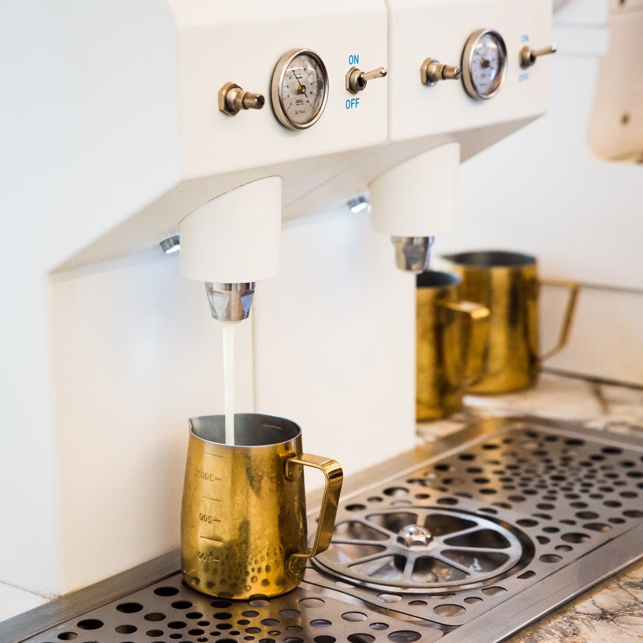 Weiße Espressomaschine füllt Milchstrahl in goldene Milchkännchen auf Edelstahl-Tropfblech, Barista-Ausstattung