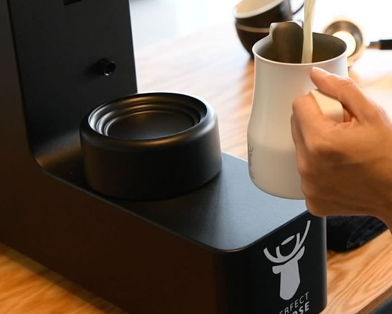 Hand hält weiße Milchkanne neben schwarzem Perfect Moose Milchaufschäumer auf Holzoberfläche