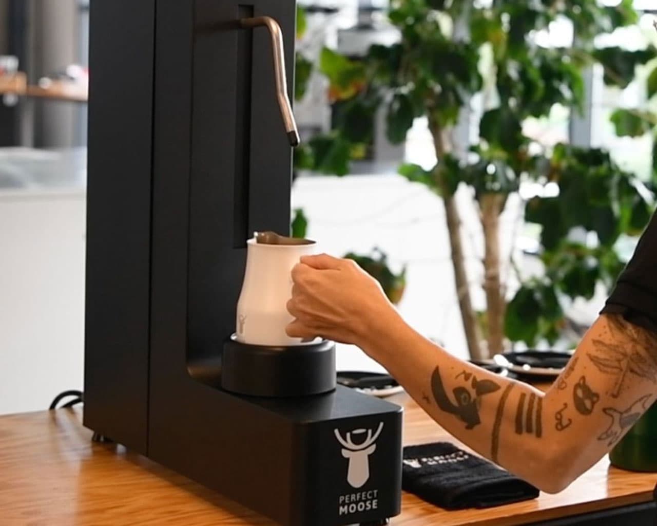 Person mit tätowiertem Arm füllt Milchkanne an schwarzer Perfect Moose Milchaufschäumer-Maschine in Caféküche