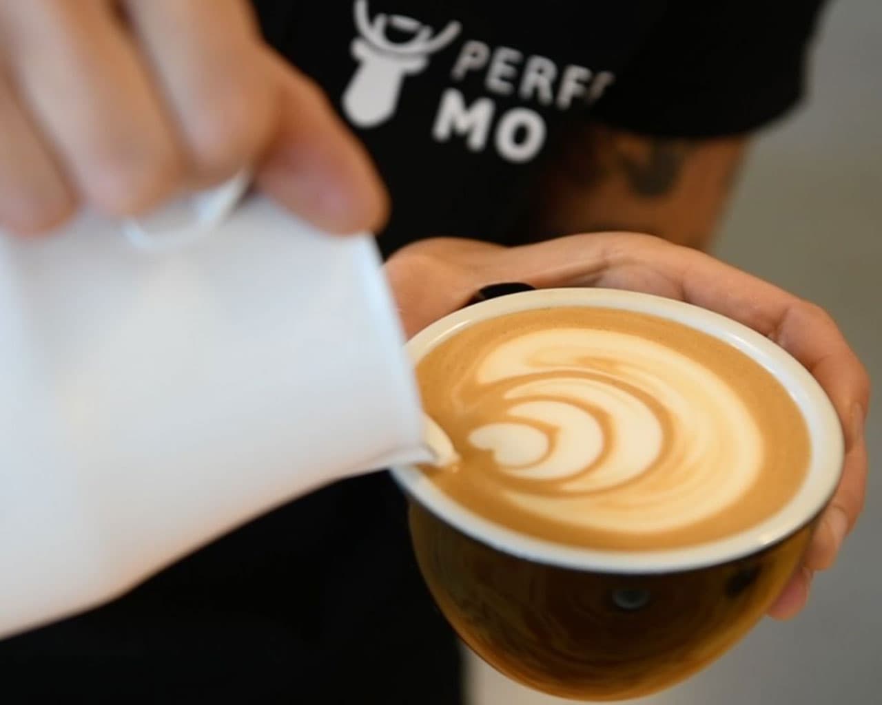 Barista gießt Milch in Cappuccino mit Latte Art, Nahaufnahme von Tasse und Hand (Perfect Moose Logo im Hintergrund)
