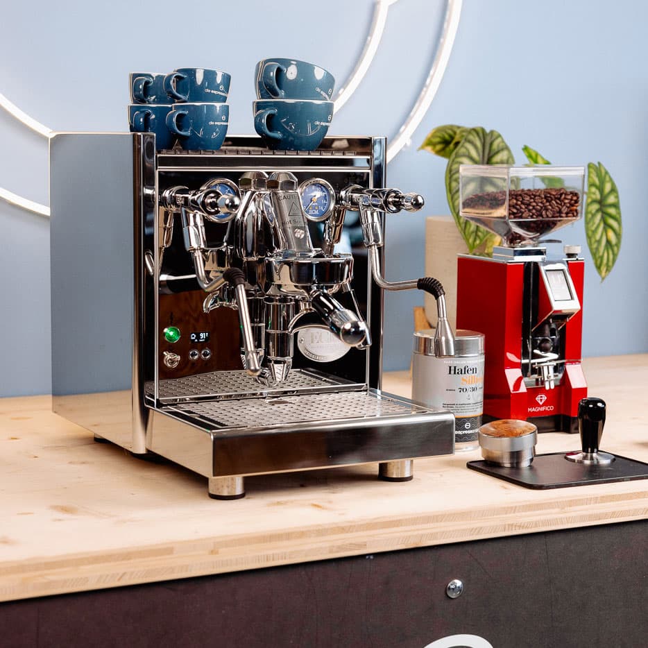 Chrom glänzende ECM Technika Siebträgermaschine mit blauen Espressotassen, roter Kaffeemühle und Zubehör auf Holzplatte
